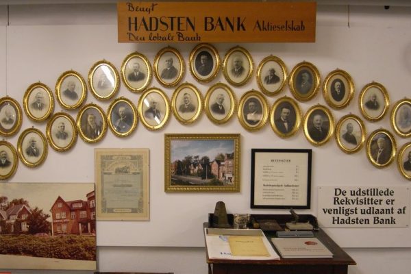 Væg med udstilling om Hadsten Bank: Ovale rammer med portrætter af direktørerne. Indrammet første aktie. Gl. rentesatser. Foto af banken m.m.